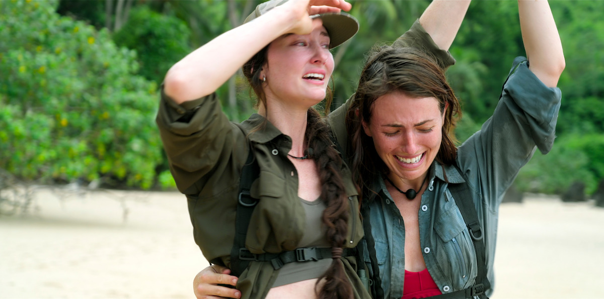 Two women in outdoor clothing, one comforting the other, both emotional on a sandy beach with lush green trees in the background, suggesting a survival or adventure scenario in a natural tropical setting.