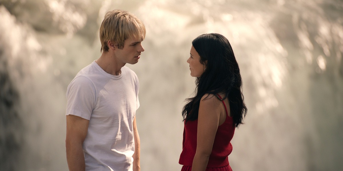 A young man in a white shirt and a young woman in a red dress stand facing each other outdoors in front of a large, flowing waterfall, engaged in a serious conversation.