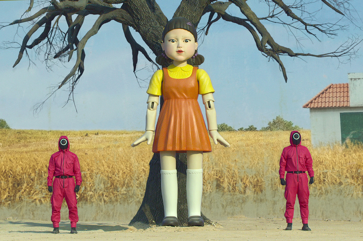  A giant doll towers over two people wearing red jumpsuits.