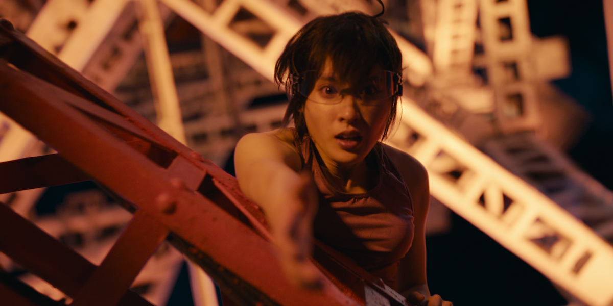 Woman in danger, reaching out while hanging from a red metal structure at night, with blurred geometric framework in the background, suggesting a tense or dramatic situation in an industrial or amusement park setting.