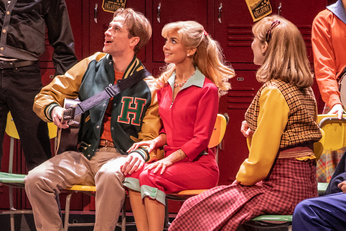 Group of young adults in 1950s-style costumes sitting on yellow chairs in front of red lockers, with one playing guitar; school musical or theater performance setting.