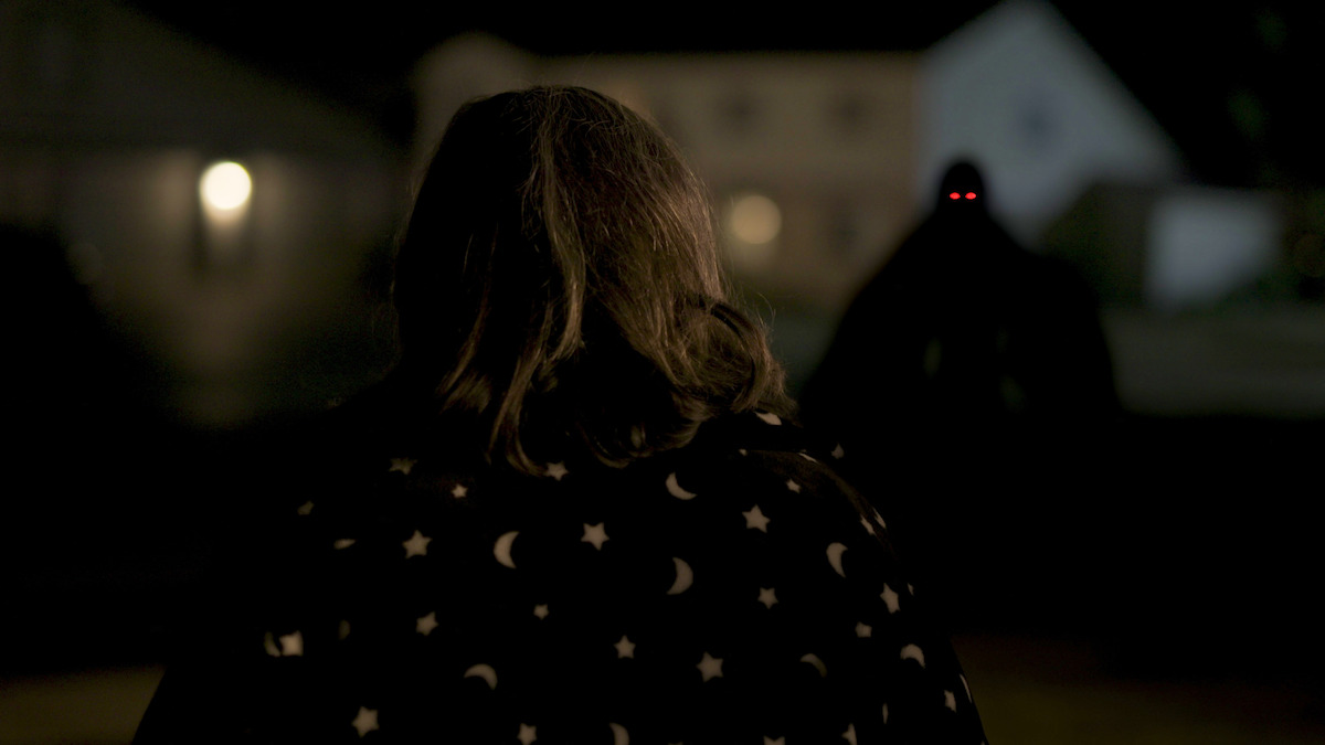 A woman stands in front of a dark, red-eyed figure.