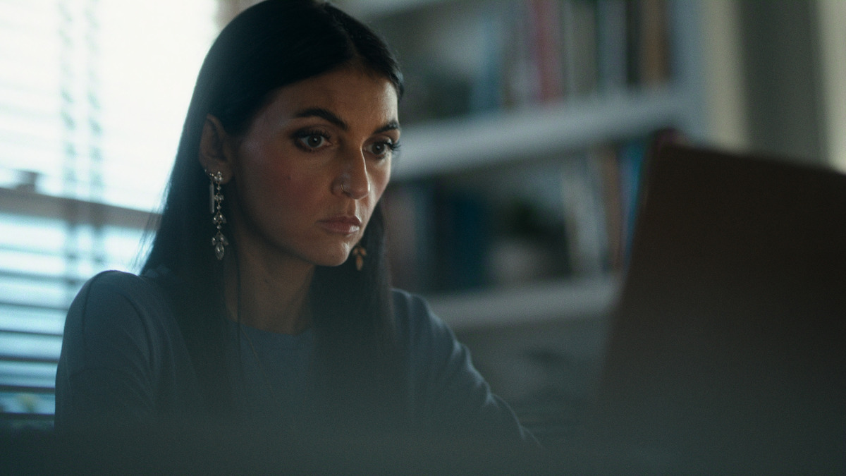 Woman with long dark hair and earrings working intently on a laptop in a softly lit room with bookshelves and a window in the background.