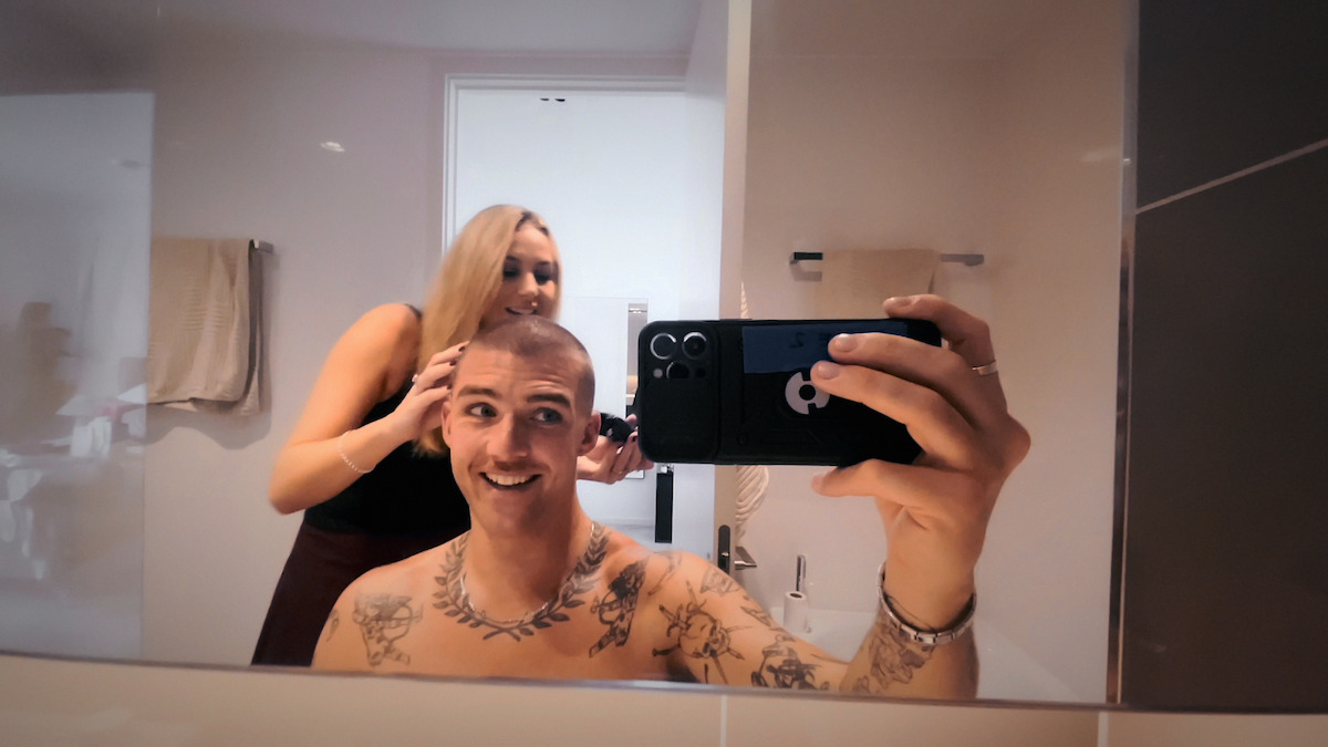 Smiling man with tattoos takes a mirror selfie while a woman stands behind him, cutting his hair in a well-lit modern bathroom with white walls and towels visible in the background.