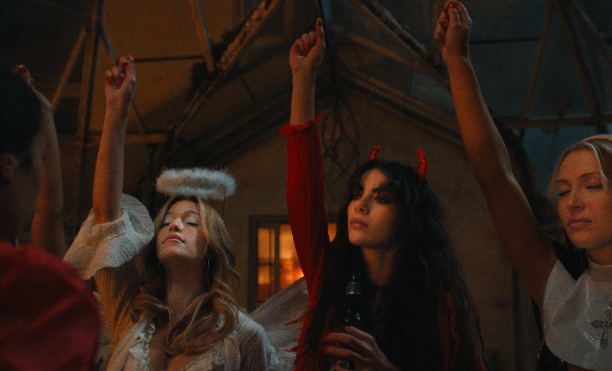 Three women in costumes at a party, one dressed as an angel and another as a devil, raising their hands in a dimly lit room with rustic decor, suggesting a festive or Halloween atmosphere.