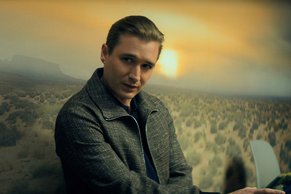 Seth Numrich as Blaine Shaw with slicked-back hair in a gray jacket sits in front of a mural depicting a desert landscape at sunset, indoors.