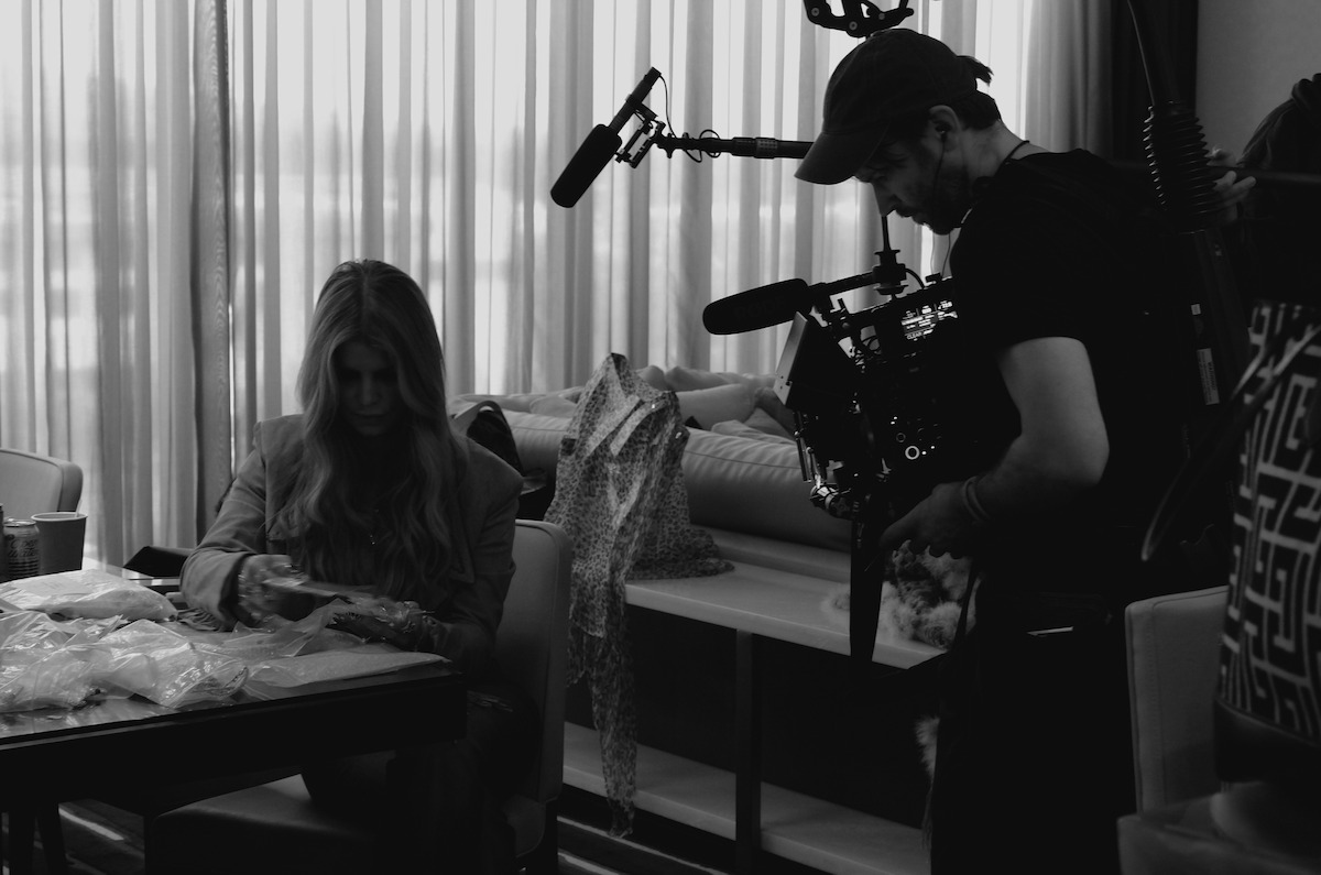 A camera operator films a woman sitting at a table indoors, with fabric samples and clothing around, in a well-lit room with large curtains in the background.