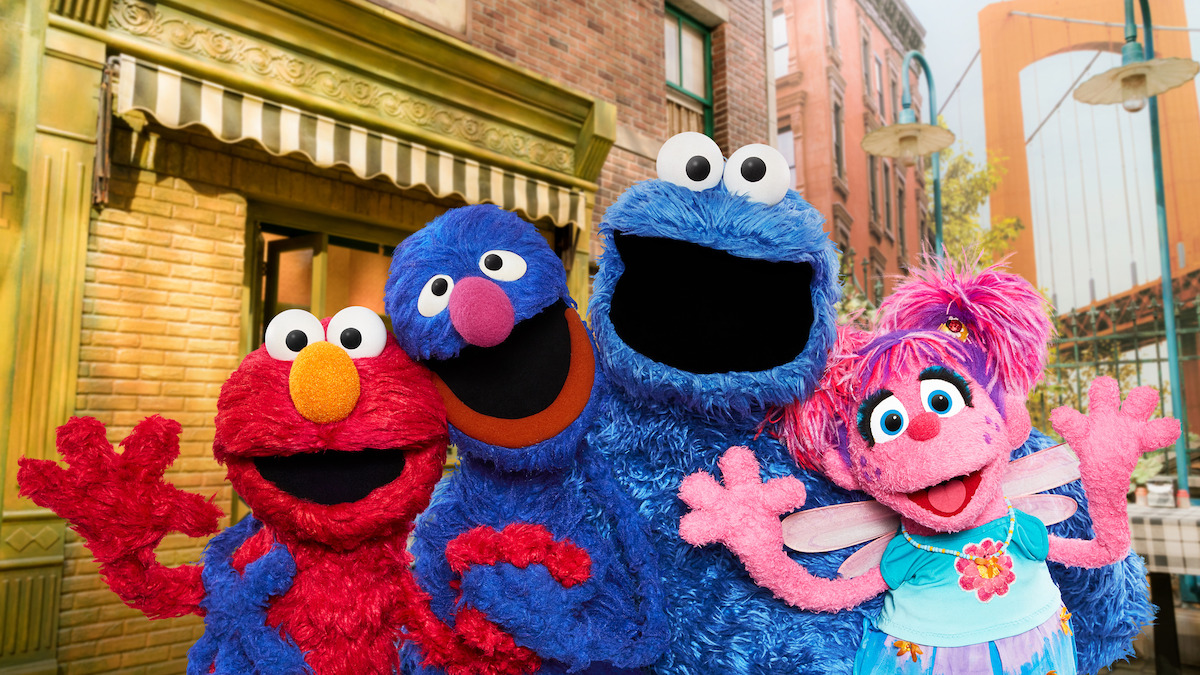 Four colorful Sesame Street puppets pose smiling together outside on a city street with brick buildings, storefronts, and streetlights in the background on a bright day.