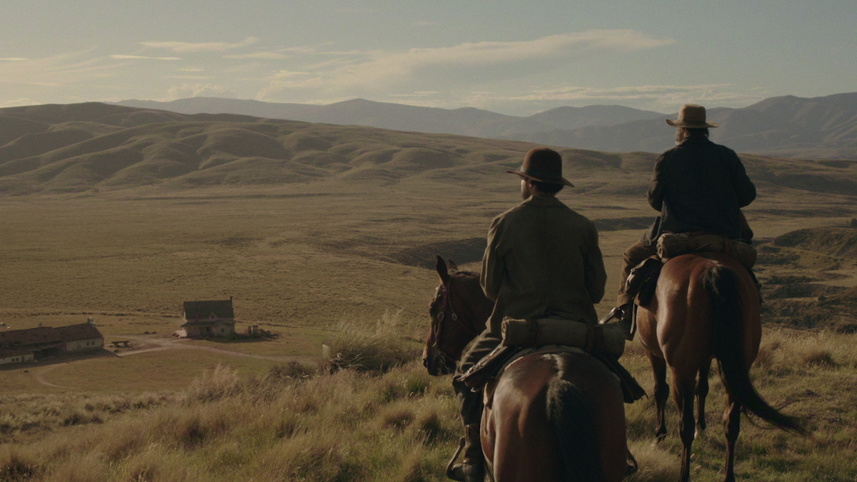 Two people on horseback overlook a wide, grassy valley with rolling hills and distant mountains, near a ranch house in the golden light of late afternoon.