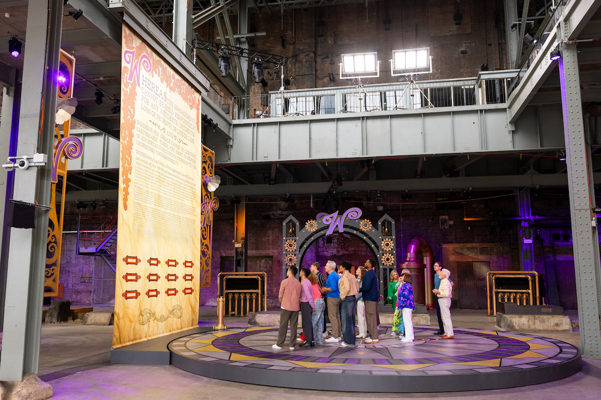 A group of people stands on a decorated circular platform inside a large, industrial-style venue with high ceilings, exposed beams, stage lights, and whimsical themed decor. A large sign and a stage with gears and a "W" logo are visible.