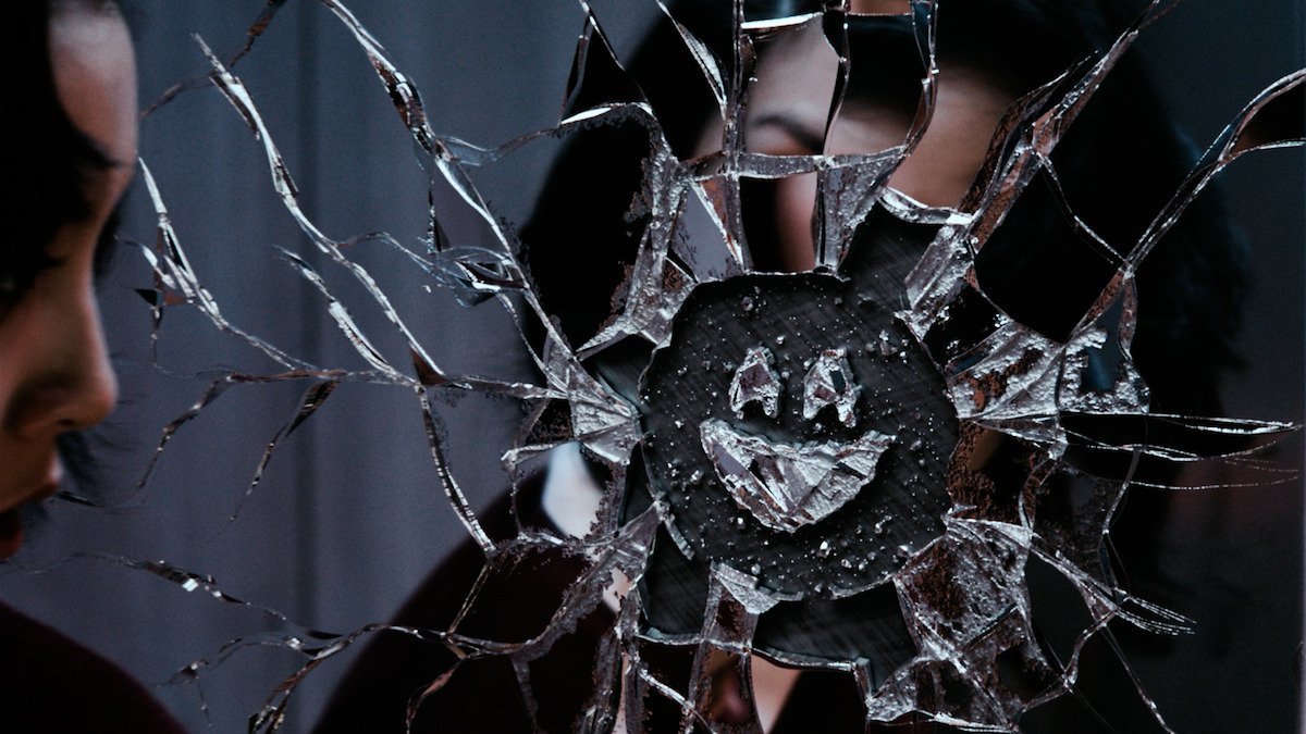 Still photo of the female character in Next on Netflix video looking into a cracked mirror featuring the ‘Black Mirror’ smiley face.