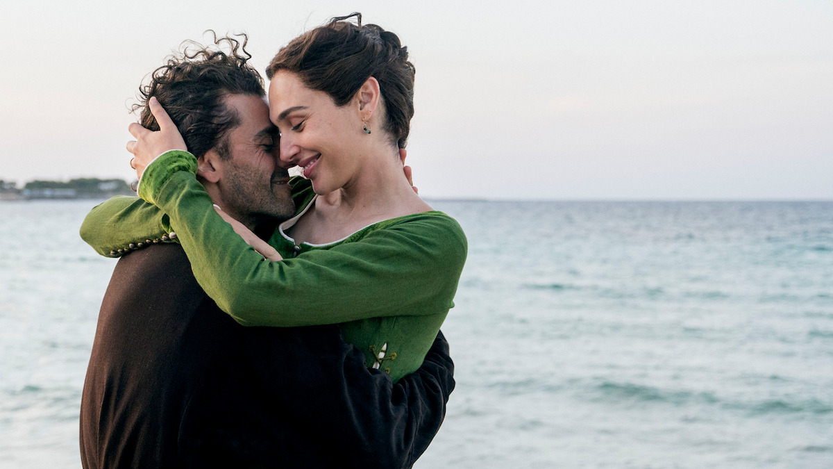 A couple embraces lovingly by the seaside, smiling at each other with the ocean and a clear sky in the background, creating a romantic and intimate atmosphere.