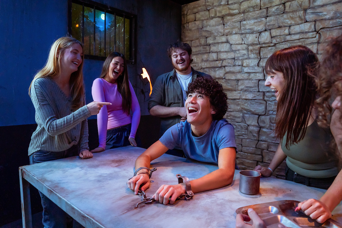 A group of young adults laughing inside a themed escape room with stone walls and dim lighting; one person sits at a metal table with handcuffs, creating a playful, immersive experience.
