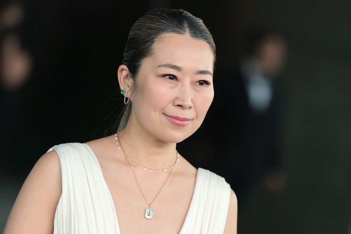A woman in a white sleeveless dress with jewelry poses with her hair pulled back. The background is dark and slightly blurred, suggesting an event or formal setting.