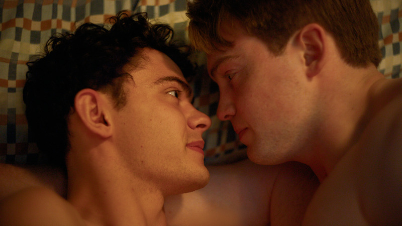 Two young men lying face to face on a bed with a checkered blanket, gazing into each other's eyes in an intimate, softly lit setting.