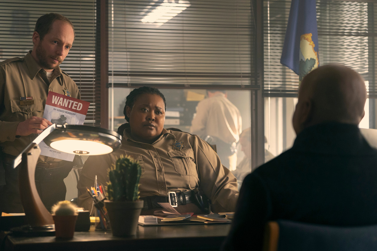 Kyle Hixon as Deputy Ryken holding a Wanted sign and Luyanda Unati Lewis-Nyawo as Detective Sinclair sitting at a desk in ‘Wednesday’ Season 2.
