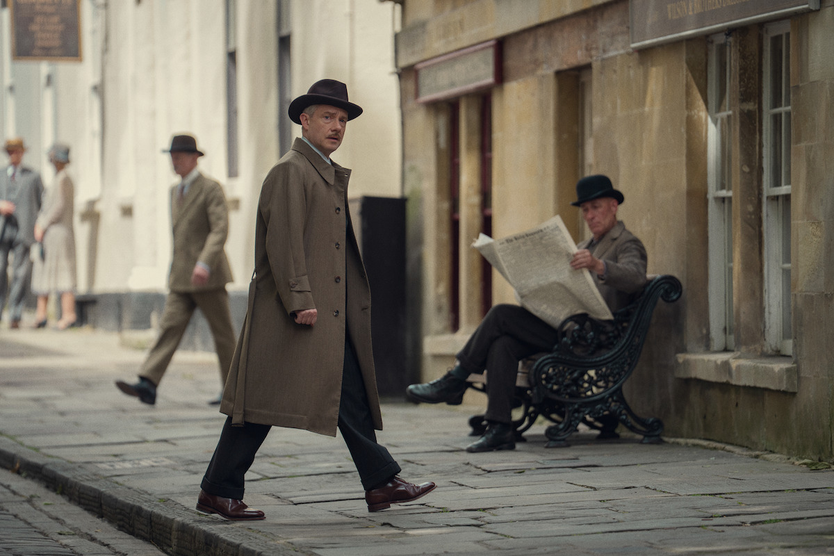 A man in a trench coat and hat walks on a cobblestone street in a historic city setting. Another man sits on a bench reading a newspaper, with two more people walking in the background, all dressed in vintage attire.