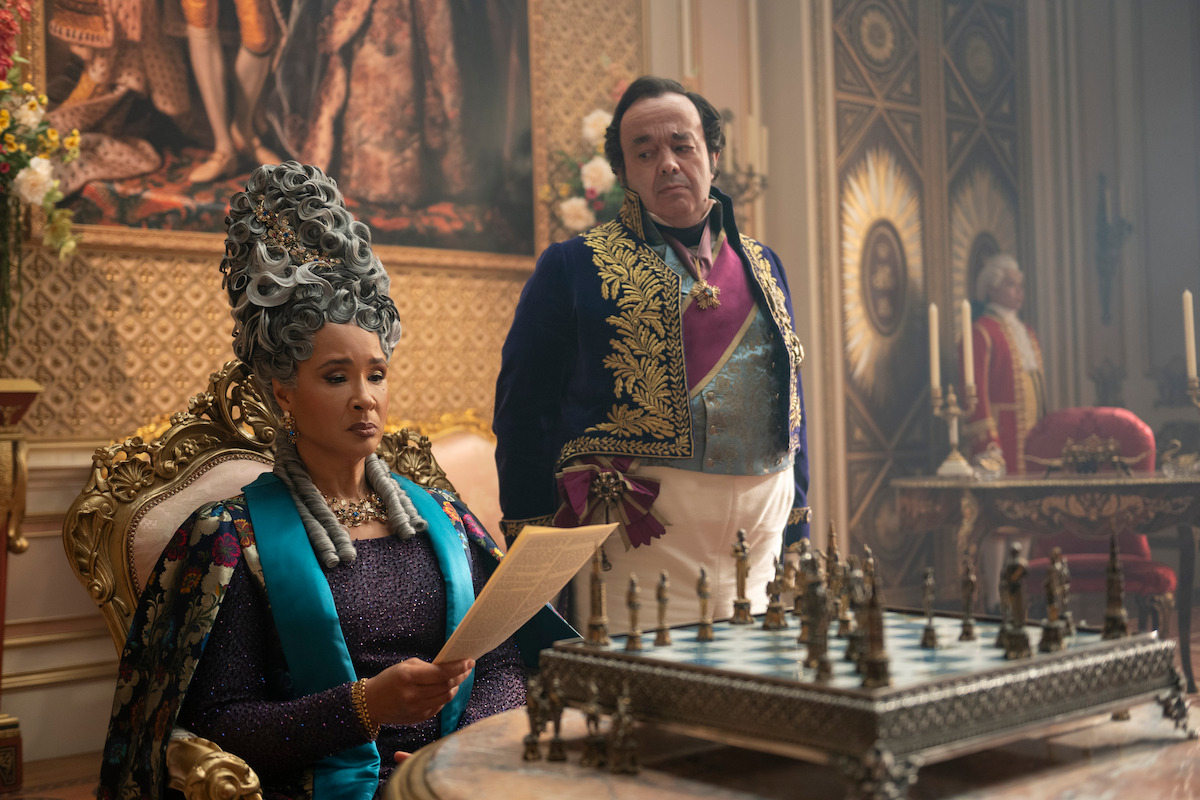A regal woman in elaborate period attire reads a letter while seated at an ornate chess table in a lavish, opulent room; a man in formal clothing stands behind her, with rich decorations and candelabras in the background.