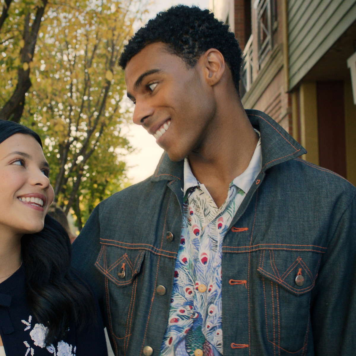 Two young adults smiling at each other while walking outdoors on a tree-lined street, with autumn leaves and buildings in the background, creating a warm and cheerful atmosphere.