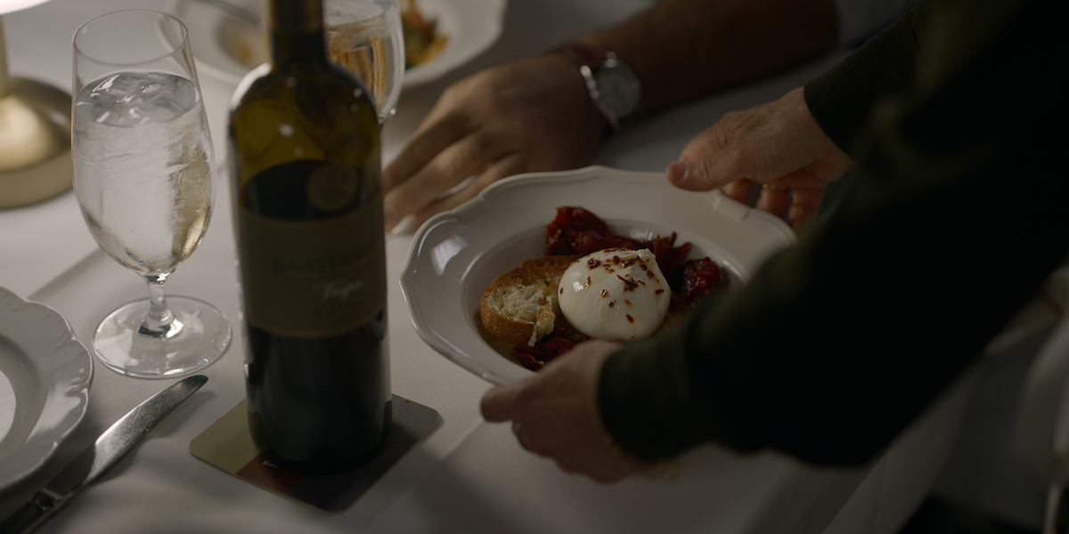 A person serves a plate of burrata cheese, bread, and tomatoes at a candlelit dining table set with wine, a glass of water, and elegant tableware, creating an intimate dinner setting.