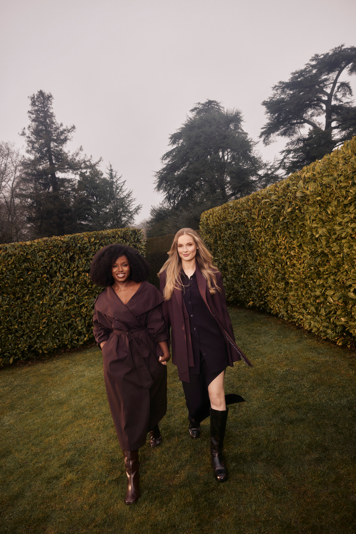 Two women walking on grass between tall hedges in an outdoor garden, both wearing dark coats and boots, with trees and cloudy sky in the background.
