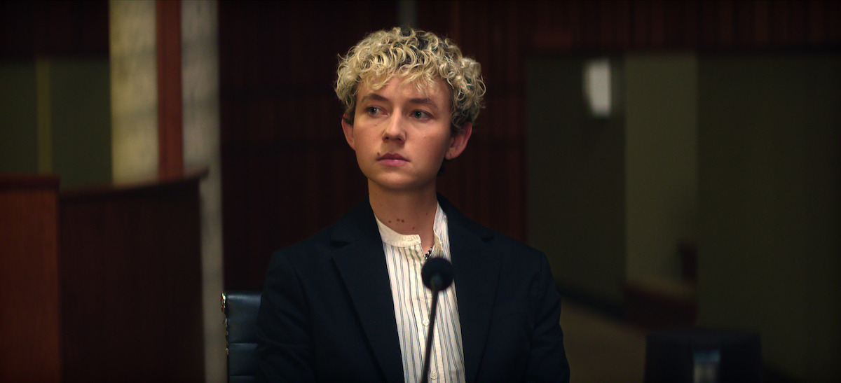 Person with short curly blonde hair wearing a suit sits at a witness stand in a courtroom, facing forward with a serious expression. Microphone in front, wooden paneling and formal setting visible in the background.