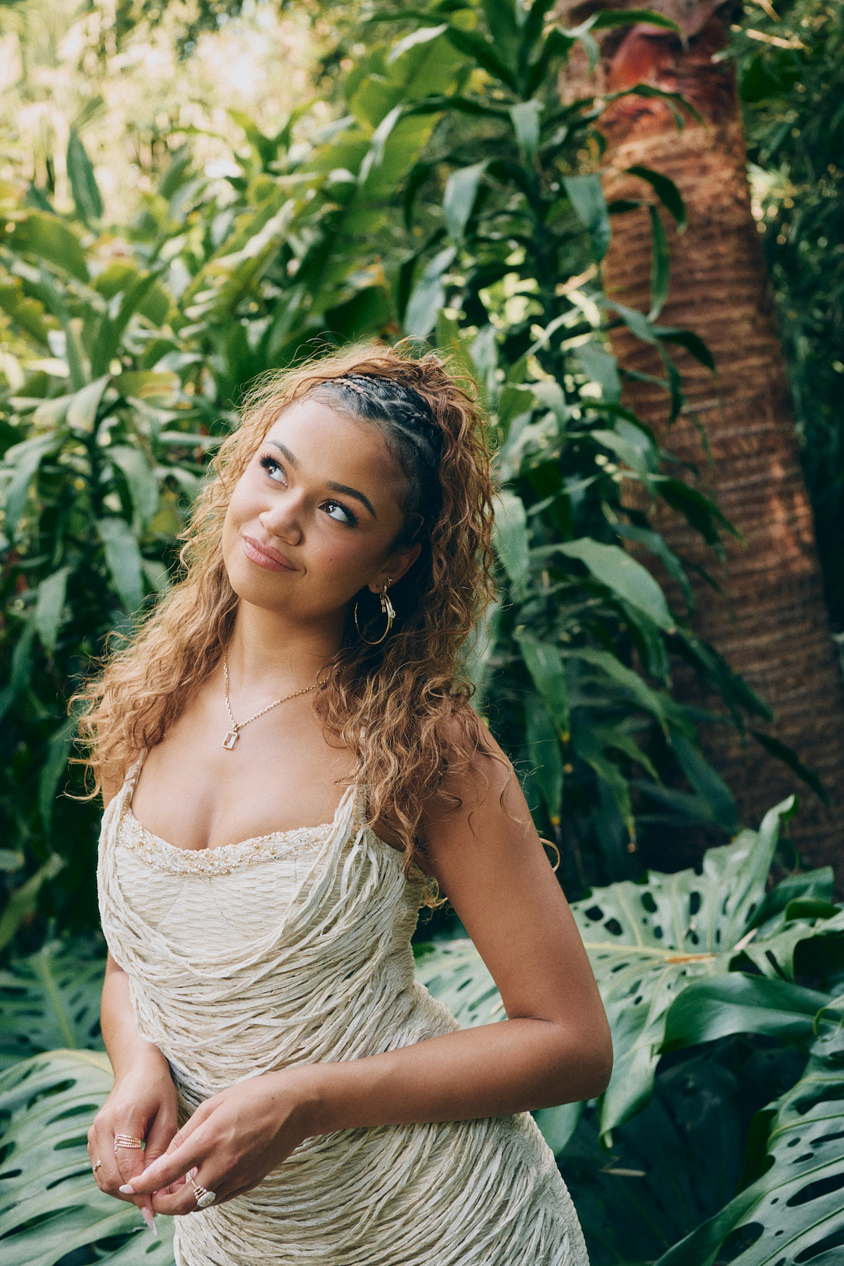 A woman with curly hair in a light-colored dress stands in a lush, tropical garden surrounded by large green leaves and trees, looking slightly upward with a thoughtful expression.
