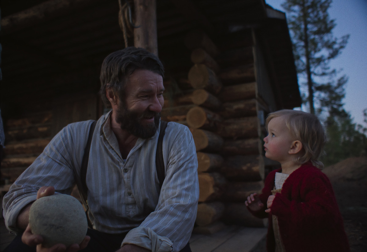 A bearded man and a young child share a warm moment outside a rustic log cabin at dusk, surrounded by nature. The man holds a ball, and both appear happy and relaxed, evoking a sense of family and tranquility in a rural setting.