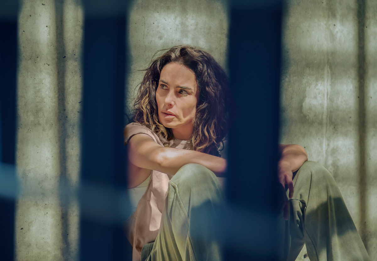 Ana Serradilla as Perla sits on the floor behind bars against a concrete wall in an image from the series ‘Sisters’ Feud.’