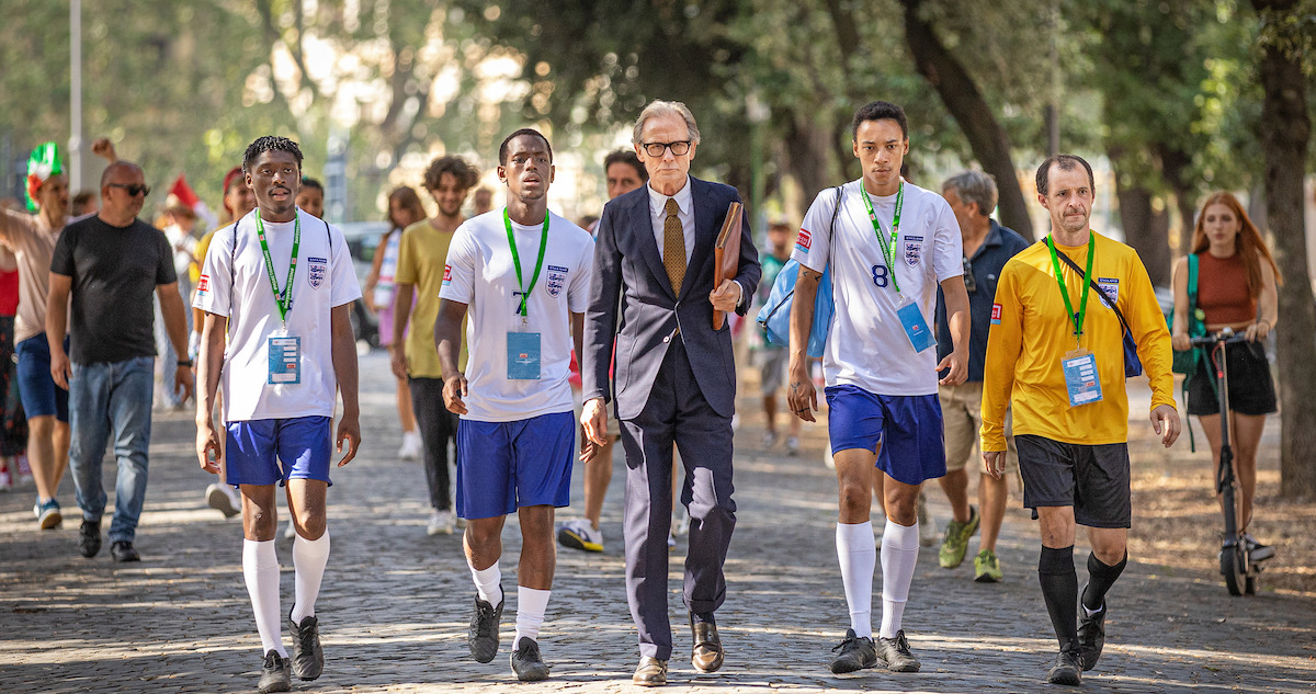 Man in suit walks alongside soccer players down a pedestrian path outside.