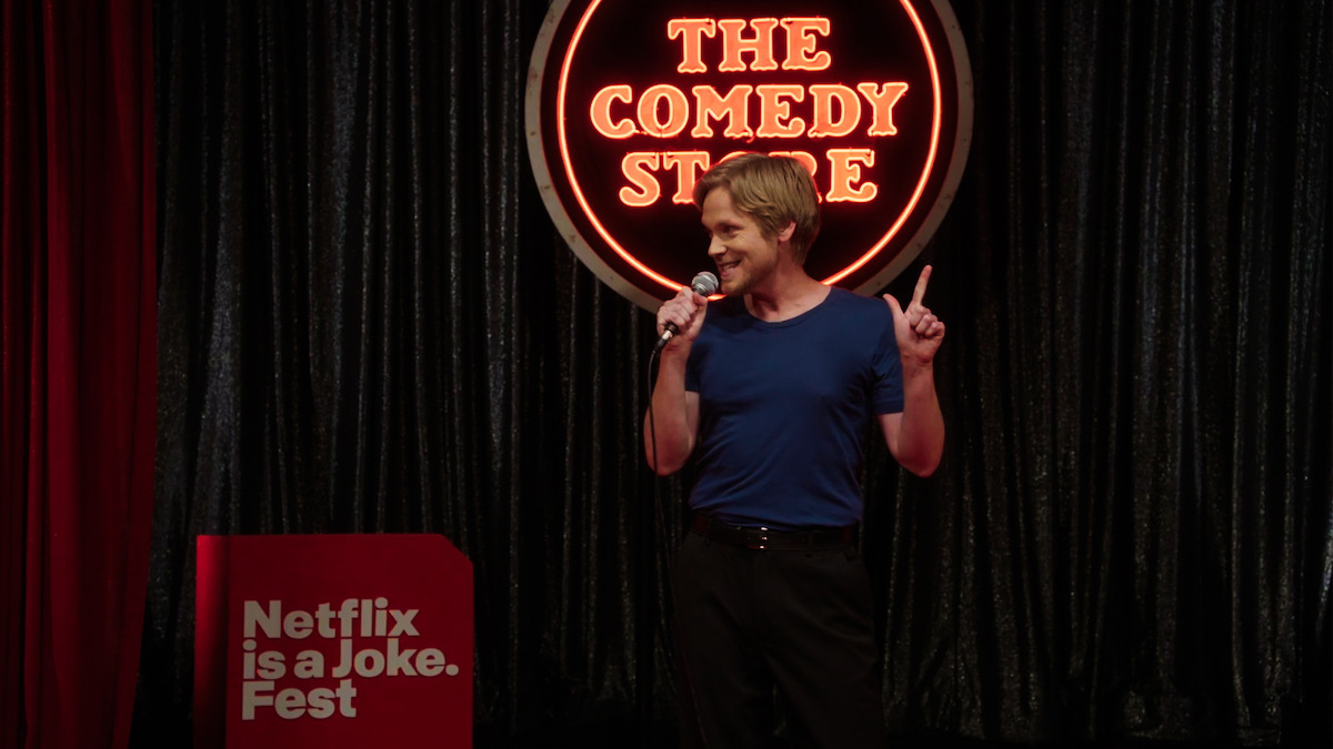 Comedian performing stand-up on stage at The Comedy Store during Netflix is a Joke Fest, holding a microphone and gesturing, with a neon sign and dark curtains in the background.
