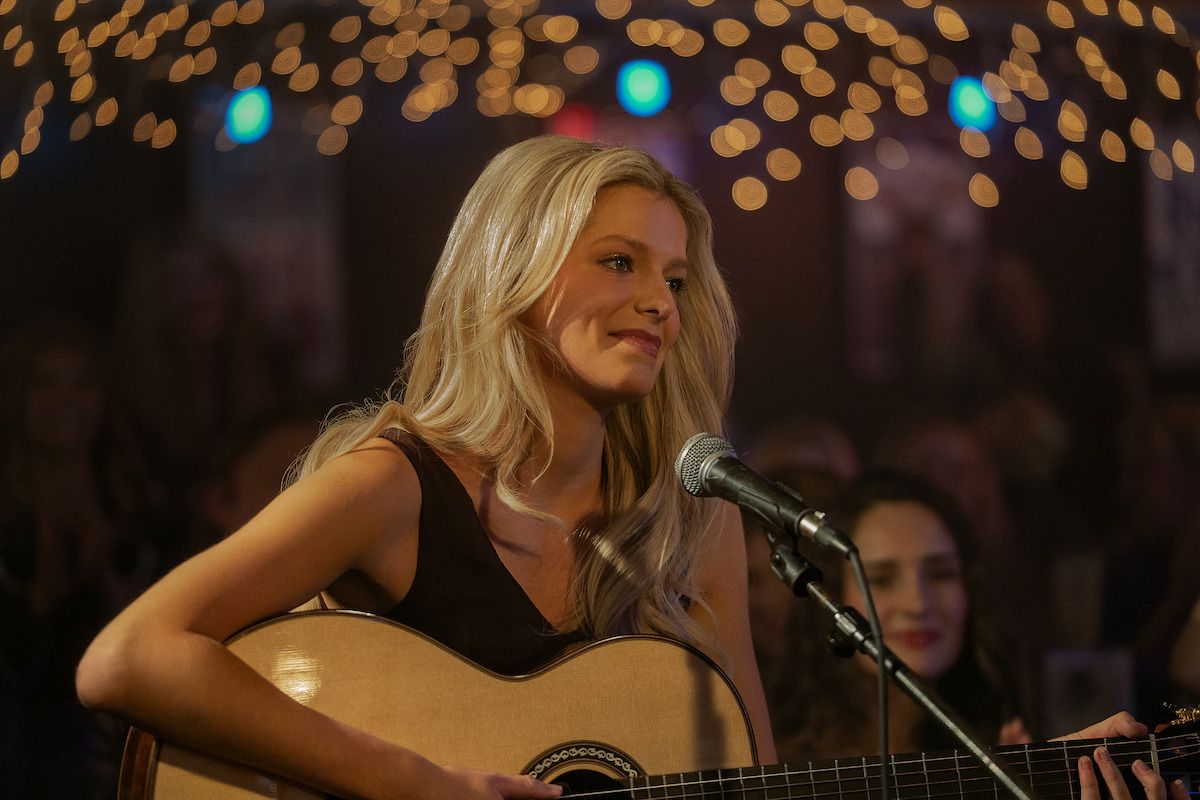 A woman with long blonde hair plays an acoustic guitar and sings into a microphone on stage under string lights, surrounded by an audience in a dimly lit, cozy music venue.