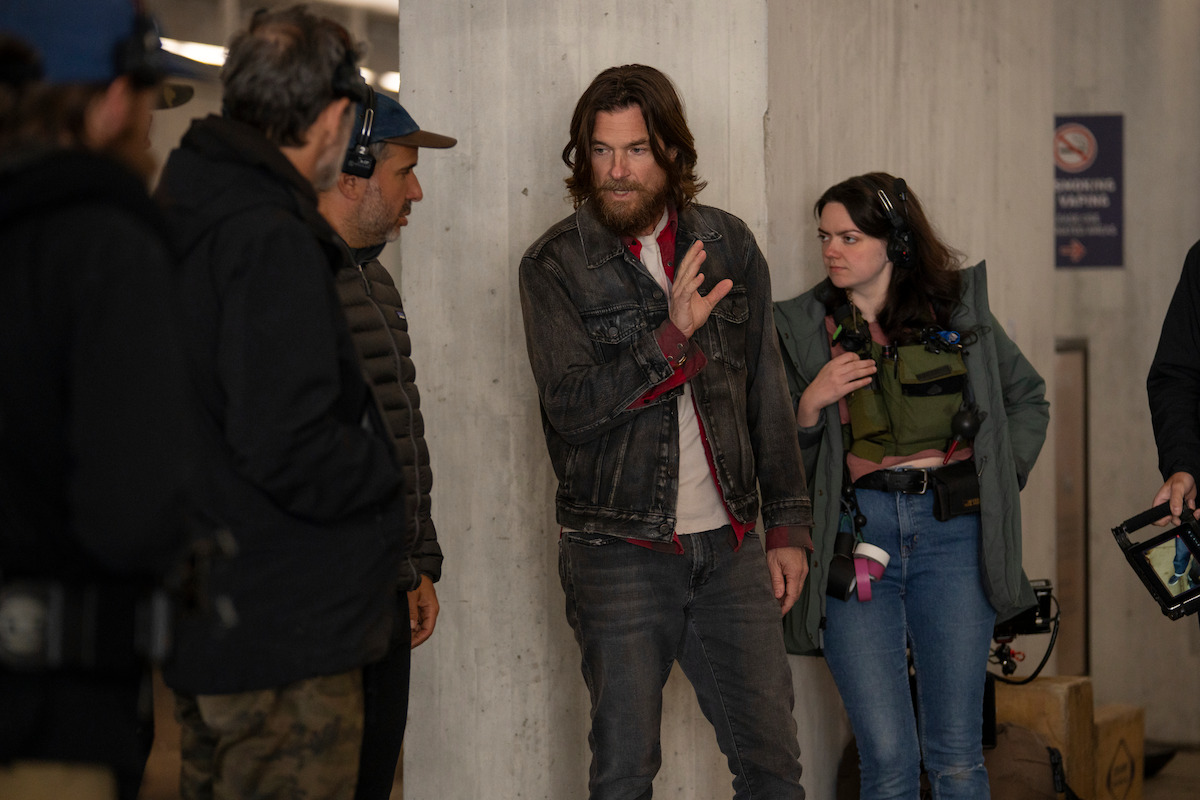 A group of people stand together indoors by a concrete wall, some wearing headsets with one man gesturing while talking, likely on a film set or during a production discussion.