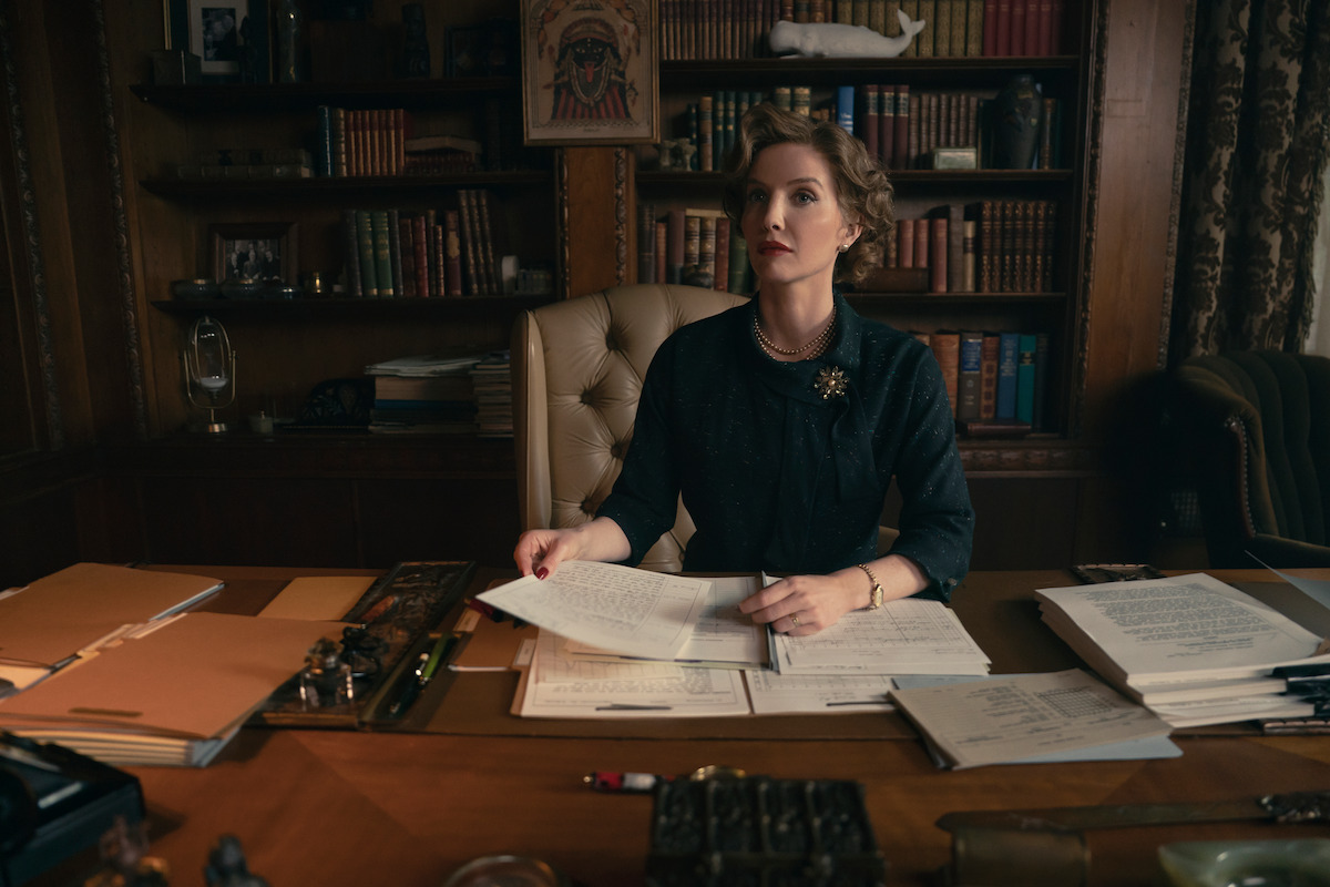 Woman sitting at a desk covered with papers and folders in a classic, wood-paneled office, with bookshelves, ornate decor, and a thoughtful, focused expression.