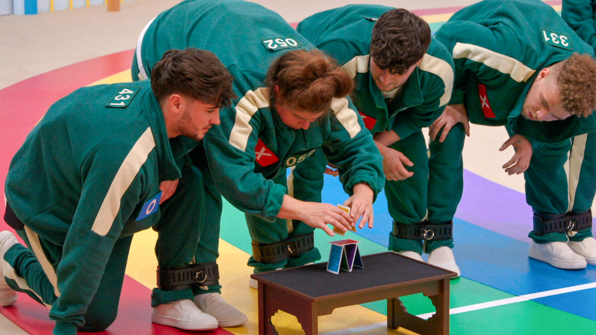 Four people in green tracksuits and numbered tags concentrate on building a house of cards on a small table in a colorful, indoor setting, suggesting a tense but playful competitive atmosphere.