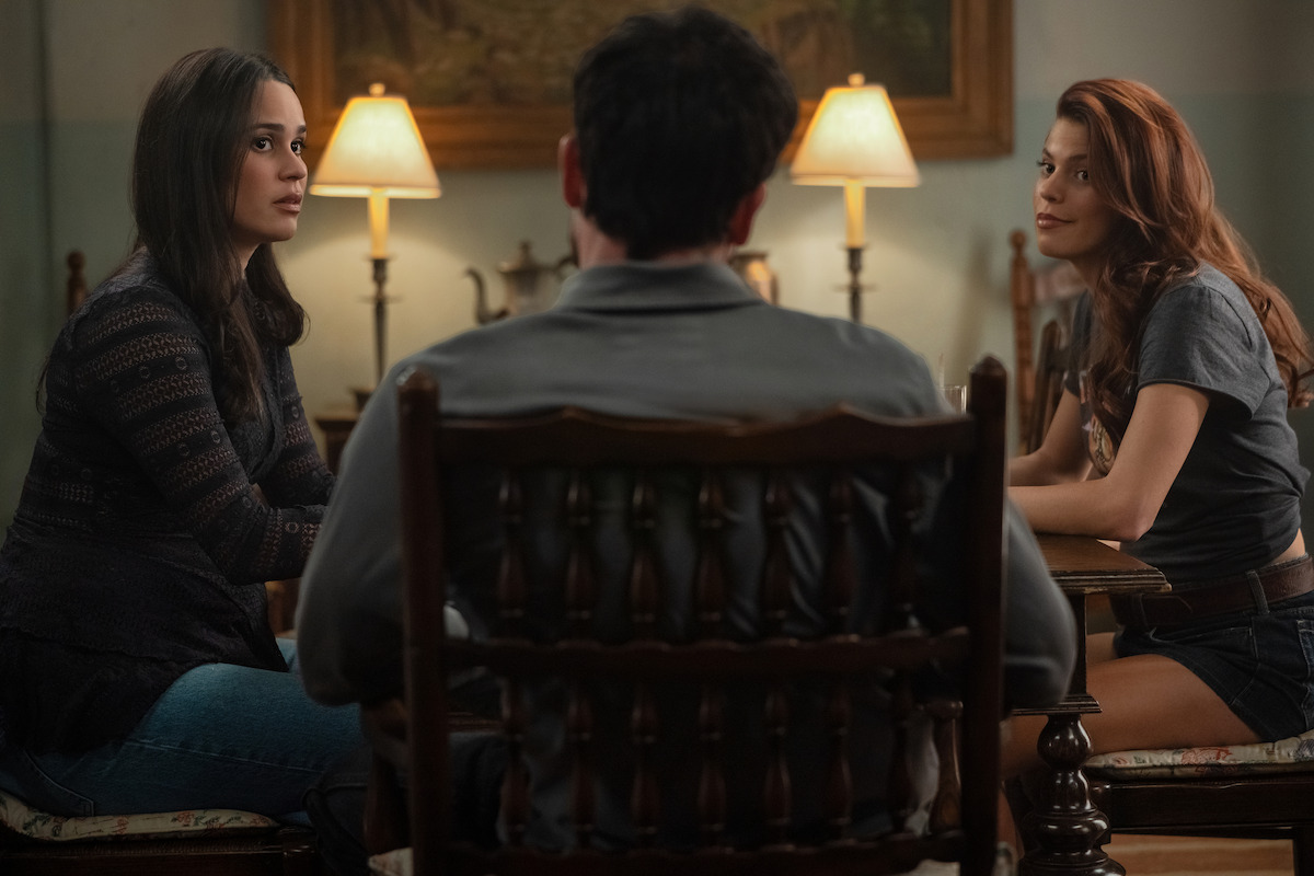 Three people sit around a table in a cozy, warmly lit room with lamps, wooden furniture, and a painting on the wall. Two women face the camera, one with long dark hair and one with reddish hair, while a man sits with back to camera.