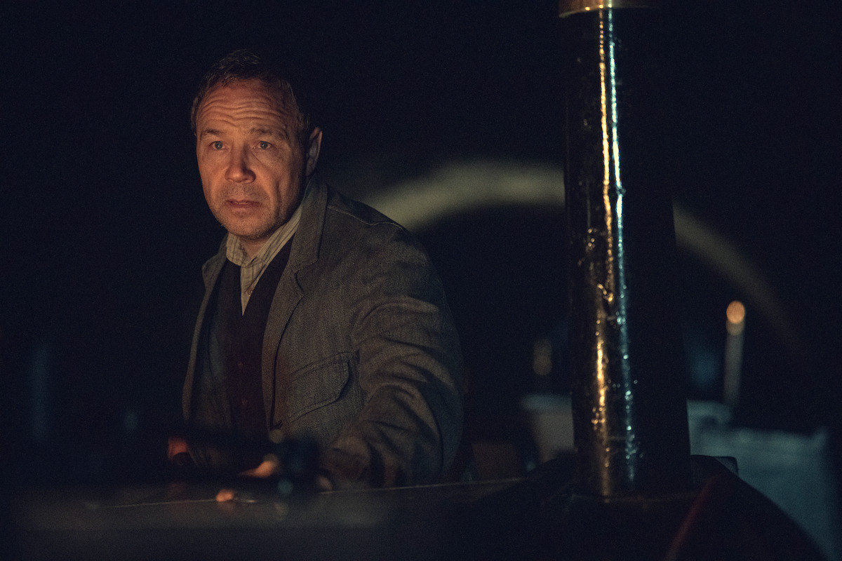 A man in a gray jacket sits in a dimly lit, dark environment, possibly inside a vehicle or industrial setting, with shadows and metal structures around him.