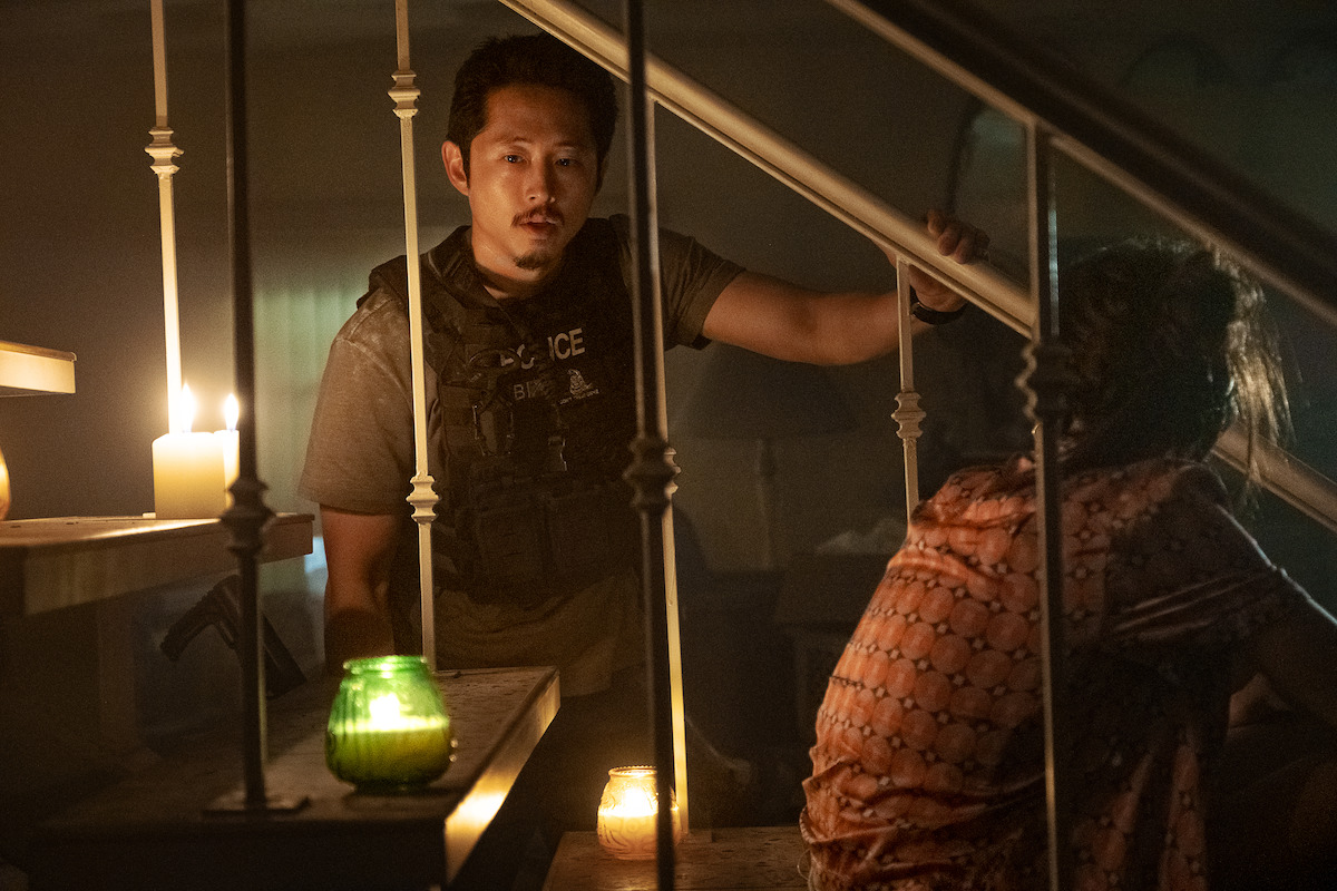 A man in a tactical vest stands by a staircase lit by candles, looking concerned, while a seated person with messy hair sits nearby in dim, moody lighting inside a house.