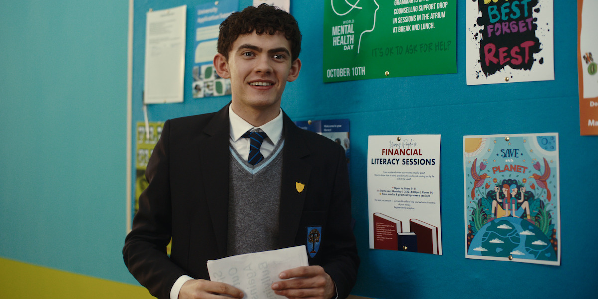 A student in a school uniform stands indoors holding papers, smiling near a bulletin board covered with colorful posters about mental health, financial literacy, and environmental awareness.