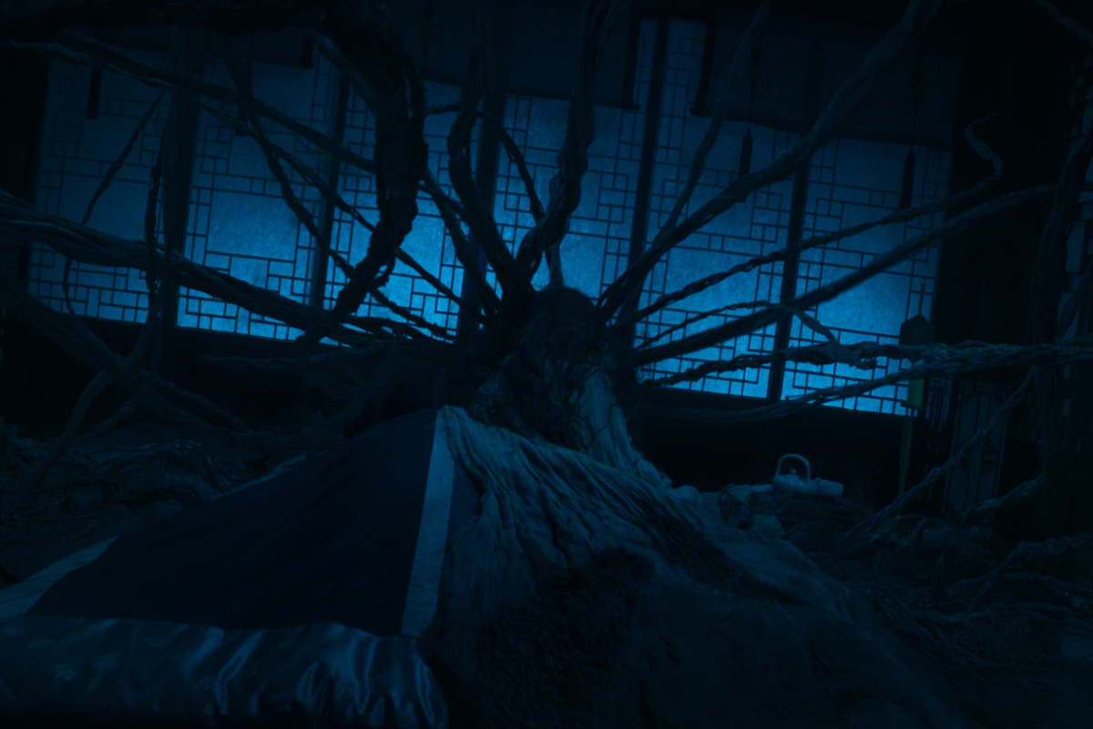 Dark, eerie room with blue lighting, showing large twisted tree roots spreading across the floor and walls, overtaking the environment. Fabric drapes over an object in the foreground, traditional window panels visible in the background.