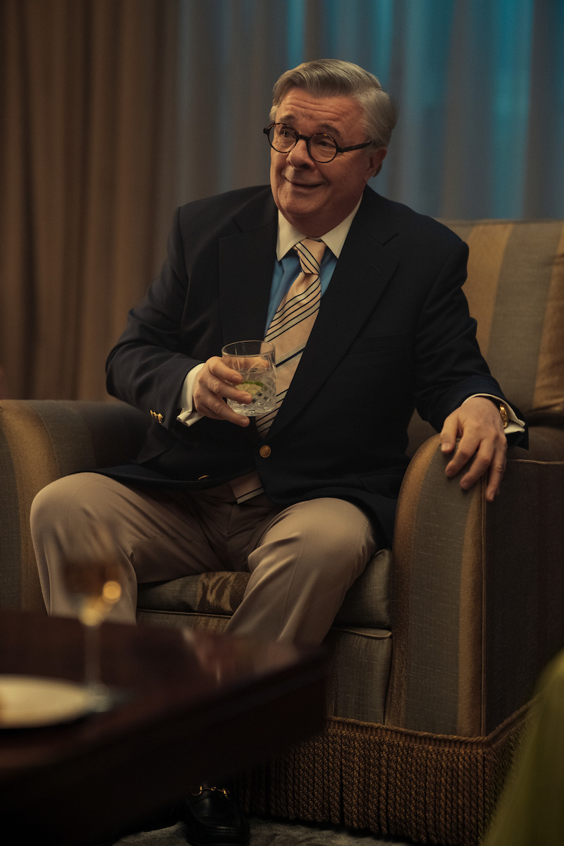 Dominick Dunne (Nathan Lane) wears a suit, turquoise shirt, and yellow tie and sits in a striped, slouchy chair.