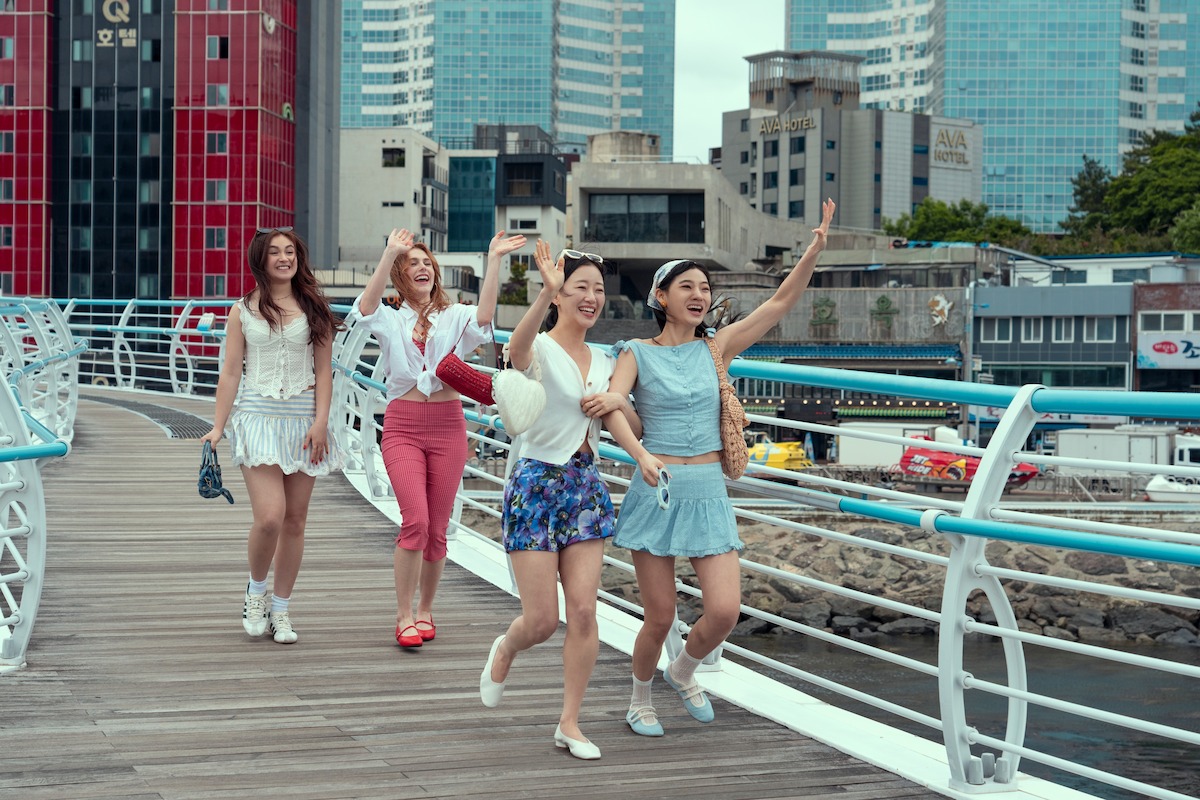 Four young women walking on a modern city bridge, smiling and waving, with tall buildings, urban structures, and a waterfront in the background on a partly cloudy day.