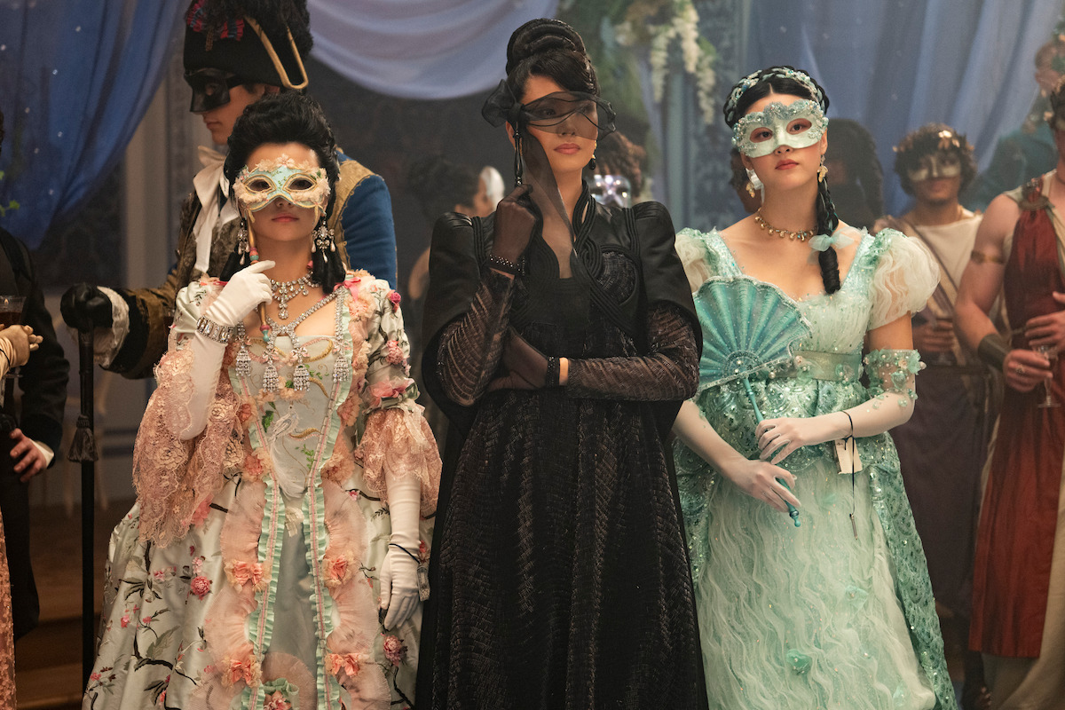 Three women in elegant, elaborate gowns and masquerade masks stand side by side at a lavish masked ball, surrounded by other masked guests in ornate costumes under draped fabric and soft lighting.