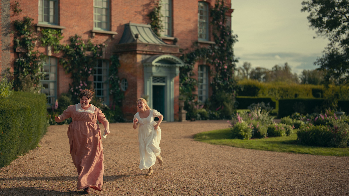 Two women in period dresses run joyfully on a gravel path in front of a large brick house with ivy and flowers, surrounded by hedges and gardens under a partly cloudy sky.