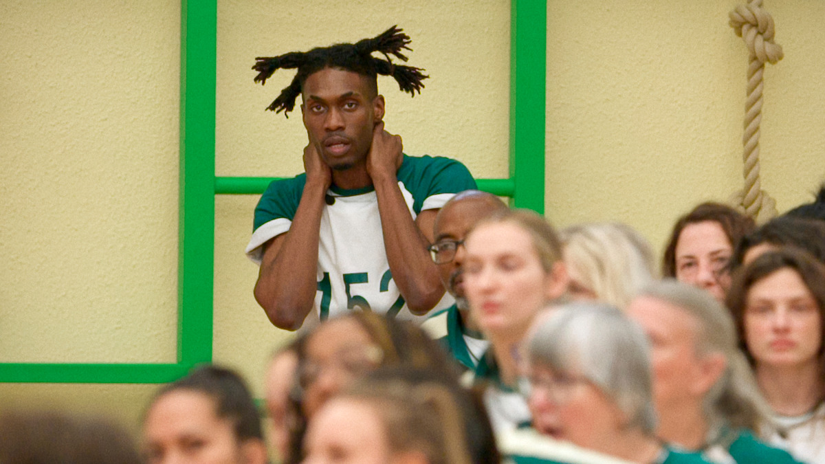 A man in a green and white jersey stands against a yellow wall and green bars, holding his head and looking tense among a crowd of people, suggesting a mood of anxiety or anticipation in a competitive setting.