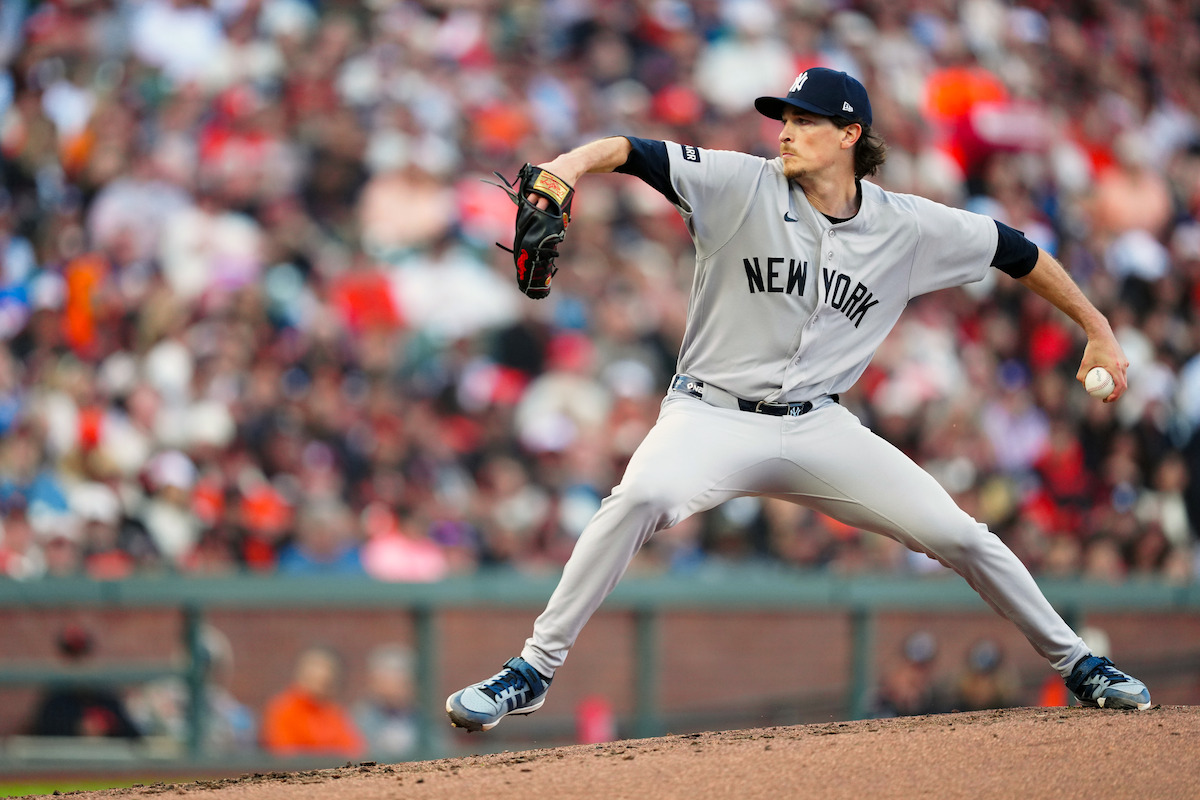 Max Fried in New York uniform winding up to throw a pitch on the mound during a game, with a blurred crowd in the stadium background.