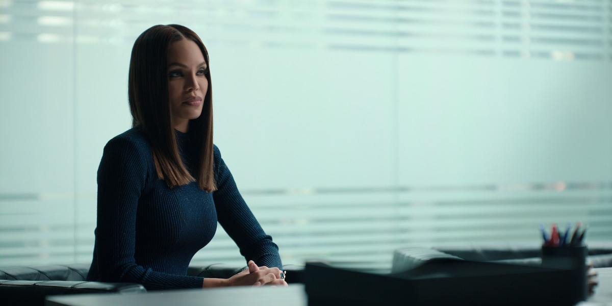 Crystle Stewart as Mallory in a dark sweater sits at a desk in a modern office with frosted glass panels and horizontal blinds, appearing calm and attentive, with office supplies visible in the background.
