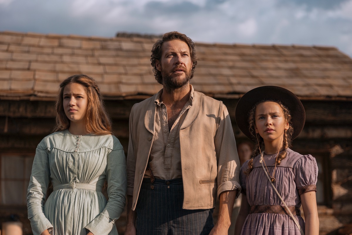 A man and two young girls in period clothing stand outside a rustic wooden cabin, looking ahead with serious expressions under a partly cloudy sky.