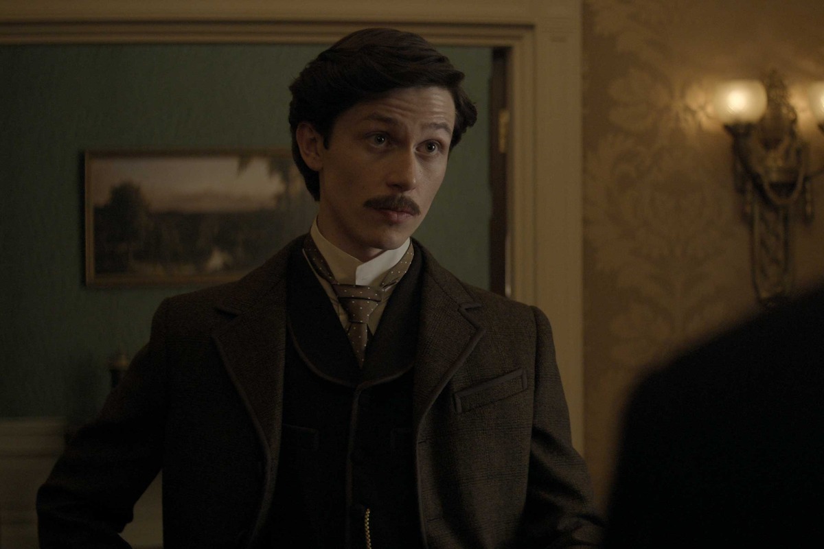 A man in vintage formal attire stands indoors in a dimly lit, ornate room, suggesting a historical setting. He appears serious and engaged in conversation, contributing to a tense, dramatic mood.