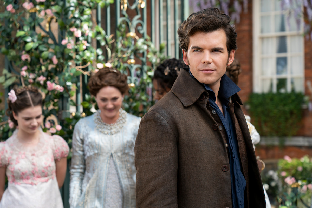 A young man in period clothing stands outdoors in front of three women in Regency-era dresses, with flowers, ironwork, and a brick building in the background, suggesting a historical or romantic setting.